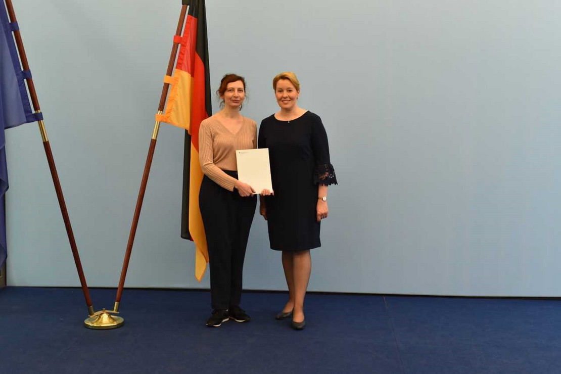Two people are standing side by side in the center next to two flags (one of Europe, one of Germany) on the left. They are standing on a dark blue carpet in front of a light-blue background. The person on the left is Claude Draude, the person on the right is Franziska Giffey.