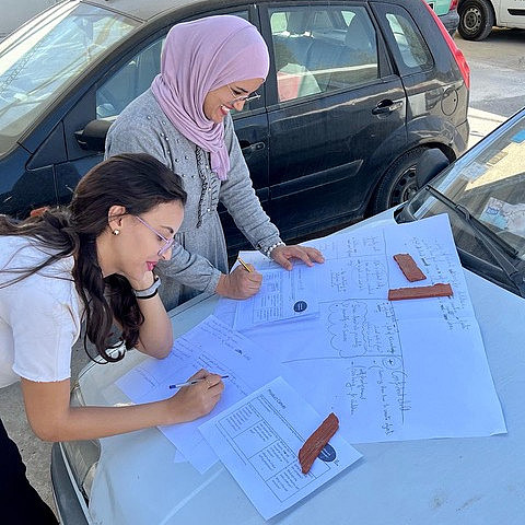 Zwei Studierende notieren etwas handschriftlich auf mehreren Seiten Papier. Diese liegen auf der Motorhaube eines Autos.