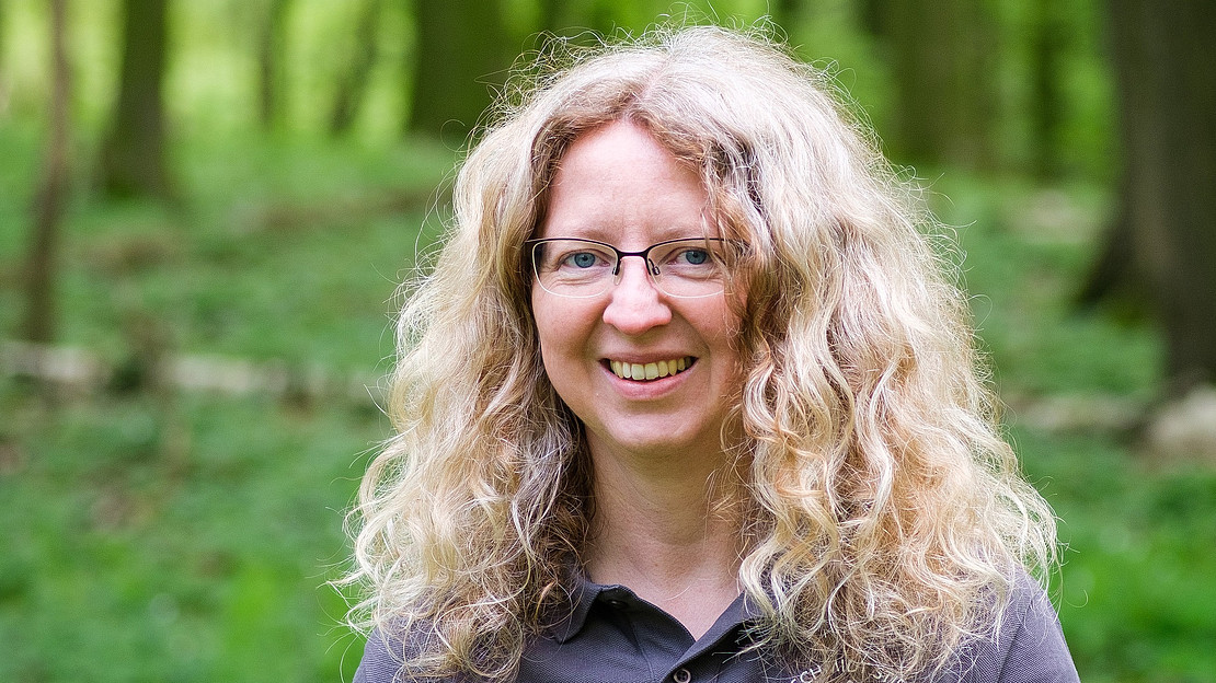 Portraitaufnahme von Kristin Luedewig. Lächelnd, lange blonde Locken, Brille, grüner Wald im Hintergrund