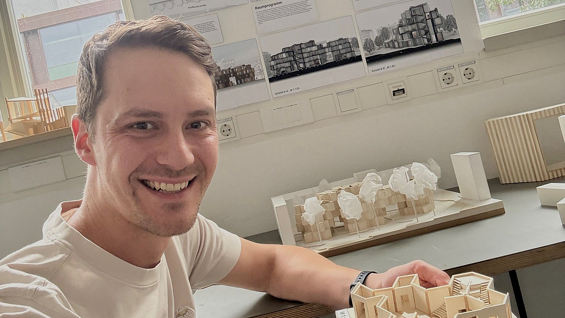 Selfie eines lächelnden Mannes am Arbeitsplatz, vor sich ein Architekturmodell