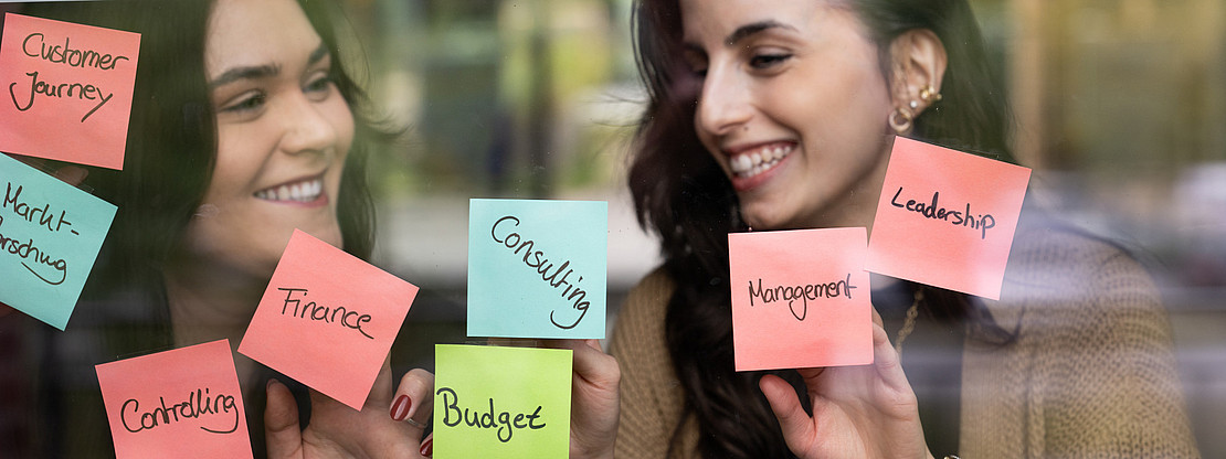 Studentinnen pinnen Wirtschaftsbegriffe an eine Glaswand.