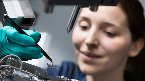  (öffnet Vergrößerung des Bildes)Das Bild zeigt eine Frau im Labor, die mit einer Pinzette eine kleine Werkstoffprobe aufnimmt, um diese zu untersuchen.