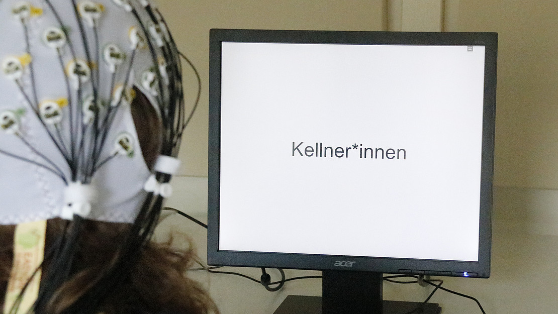 Das Bild zeigt eine Person vor einem Computerbildschirm: Mit am Kopf angebrachten Elektroden wurde die elektrische Aktivität des Gehirns beim Lesen verschiedener Satzkombinationen gemessen